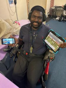 A smiling man in a wheelchair holds a tablet and a smartphone, surrounded by a casual indoor setting.