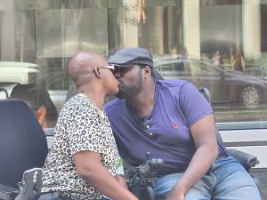 A couple shares a kiss while sitting outside, with a city backdrop visible behind them.