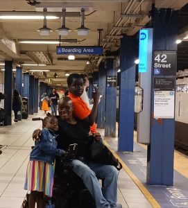 A family poses at a subway station, with a woman in a wheelchair and two children, one wearing a colorful striped dress.